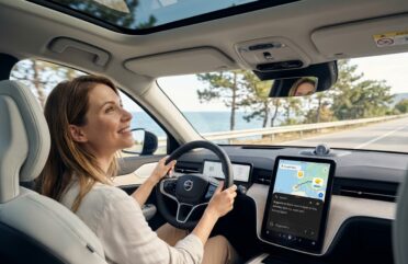 Жена шофира модерно Volvo по крайбрежен път, докато използва Google Gemini на централния екран за планиране на пътуване.